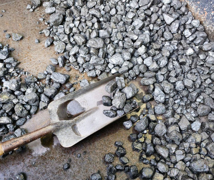 Hand holding a lump of coalA metal shovel resting on a surface scattered with shiny, black coal chunks. The coal appears freshly mined or prepared for use, with a rugged, metallic backdrop visible.