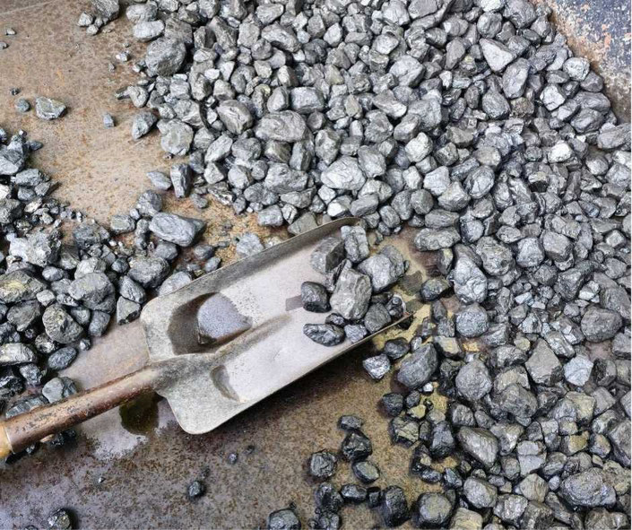 Hand holding a lump of coalA metal shovel resting on a surface scattered with shiny, black coal chunks. The coal appears freshly mined or prepared for use, with a rugged, metallic backdrop visible.
