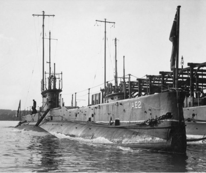 A vintage submarine marked "AE2" is docked near a wooden structure, with a flag raised at the stern and a crew member standing on its deck.