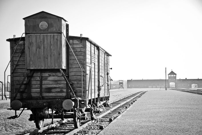 A black and white image of a wooden train car on tracks leading toward the entrance of a concentration camp with a distant watchtower.
