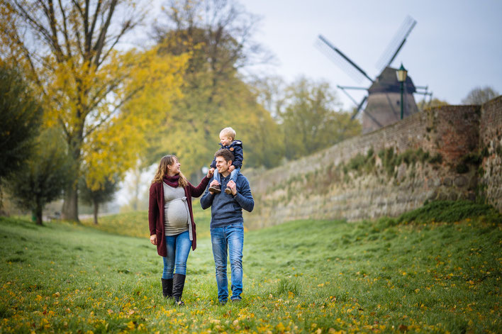 Schwangere Frau läuft mit Partner und Sohn auf der Schulter über eine grüne Wiese.