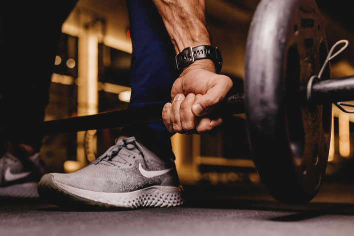 Ein Mann ist im Fitnessraum und macht Kreuzheben. Man sieht seine Hand, die die Langhantelstange greift. Die Stange befindet sich kurz über seinem Fuß.