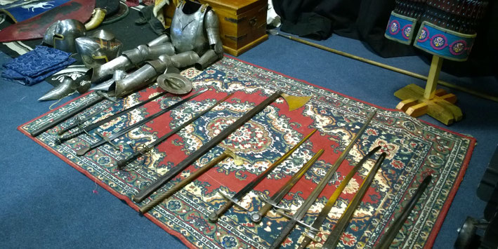 An assortment of medieval weapons and armor displayed on a patterned rug. It includes swords, polearms, helmets, gauntlets, and a breastplate, suggesting a historical reenactment setup.