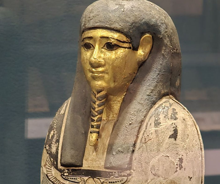 An ancient Egyptian statue of a man with a golden face, black-lined eyes, and a headdress. The worn paint and intricate details suggest it was part of a funerary or religious artifact.