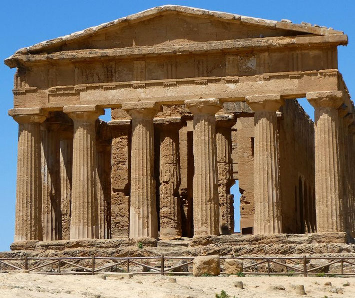 An ancient Greek temple in Sicily, likely the Temple of Concordia in Agrigento. It features well-preserved Doric columns and a pediment, standing against a bright blue sky.