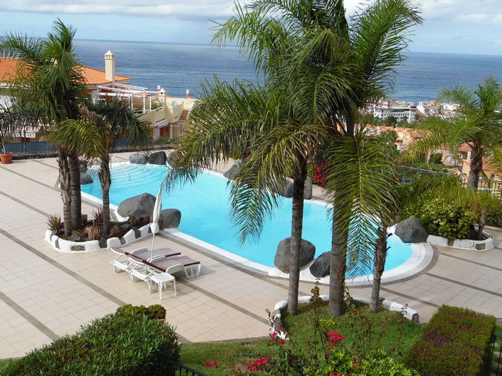 Blick auf den Pool, der mit Natursteinen und Palmen gestaltet ist, von der Wohnung aus. Im Hintergrund sieht man den Atlantik.