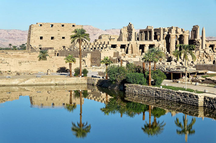 Ancient Egyptian temple ruins with palm trees reflected in a still water basin under a clear blue sky.