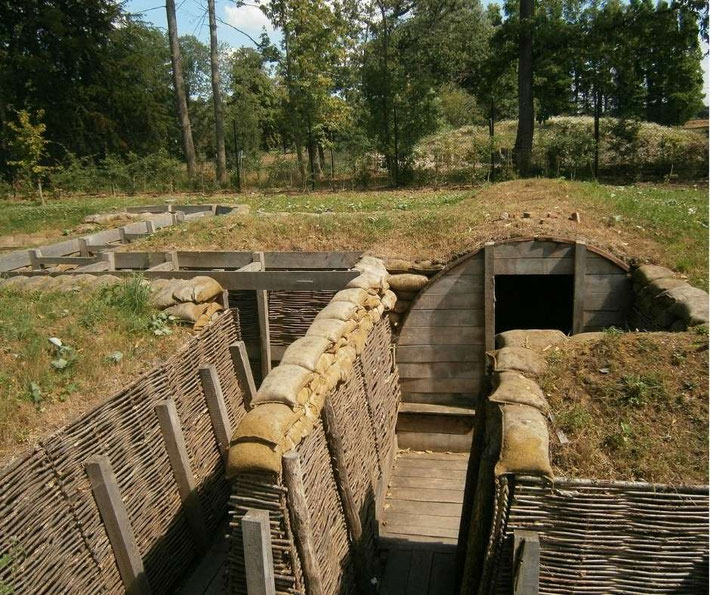 A reconstructed World War I trench with sandbags, wooden supports, and a dugout entrance, set in a grassy, wooded area.