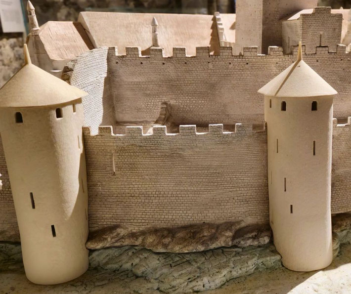 A detailed model of a medieval castle features concentric stone walls, battlements, and two cylindrical towers. The miniature design highlights historical fortifications and defensive architecture.