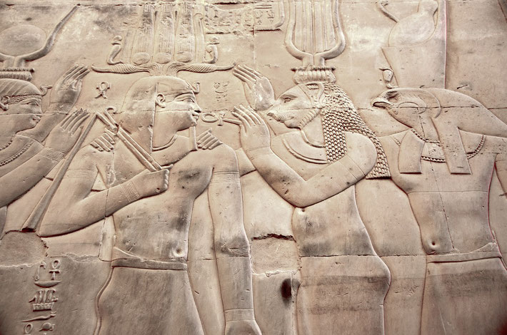 An Egyptian relief shows a pharaoh receiving an ankh from deities, including a lion-headed goddess and falcon-headed Horus. The intricate carvings depict divine blessings and protection.