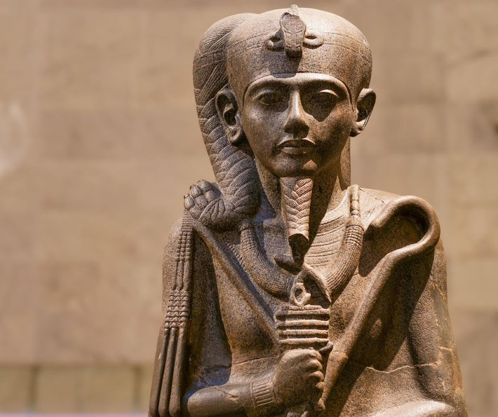 A stone statue of the ancient Egyptian god Khonsu. He wears a sidelock, a uraeus, and a false beard, holding a was-scepter.