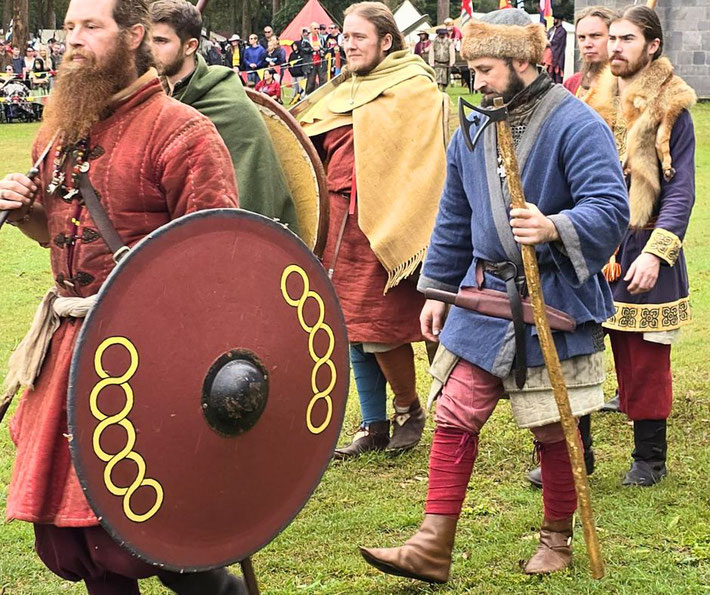 A group of historical reenactors dressed as medieval Norsemen, wearing colorful tunics, cloaks, and fur accents, carrying shields and axes, participating in what appears to be a public event.