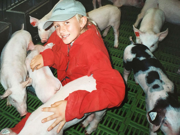 Ein blondes Mädchen, das einen roten Overall und eine Cappy trägt, sitzt in einem Stall auf der Erde zwischen Ferkeln und lächelt in die Kamera.