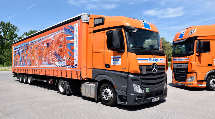 IM blauen Himmel stehen die Worte Maschinentransporte aus Leidenschaft. Man sieht weiße Wolcken und 2 LKW in orange fahren über eine Brücke. Vorne 1 kleiner, dahinter 1 LKW für Maschinentransporte.
