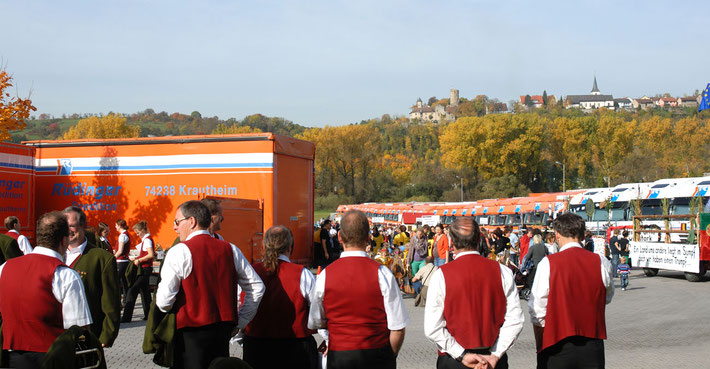 Mans ieht einen riesigen PArkplatz mit LKW für den Maschinentransport. Dazwischen sind am Besuchertag viele Menschen, im Vordegrund sieht man 6 Mitglieder einer Kapelle in rot/weiß. Der Himmel blau.
