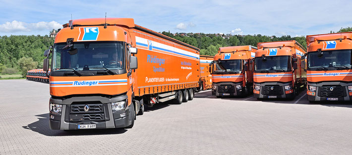 Maschinentransport aus Leidenschaft steht oben links in großen Lettern. Vor einem orangenem modernen Neubau steht ein riesiger LKW für Maschinentransporte in oragnener Farbe. Links sieht man einen kle