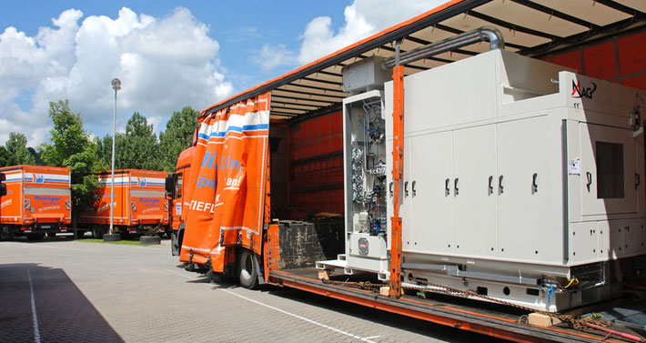 Man sieht einen Maschinentransport LKW mit 3 grünen landwirtschaftlichen Maschinen. an der Seite ist die Plane offen. Der Maschinentransporter ist orange, er steht auf dem Betriebshof.