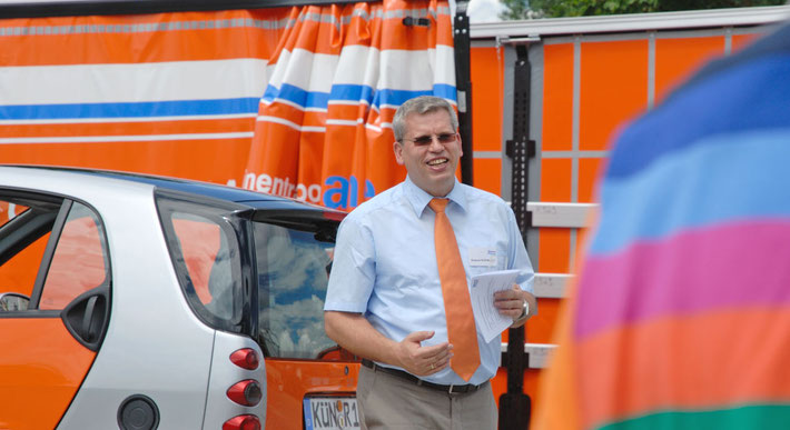 In großen weißen Lettern liest man im Bild Maschinentransporte aus Leidenschaft. Im Bild sieht man den Inhaber der Spedition, er spricht zur Kamera und steht vor 2 Fahrzeugen in Orange.