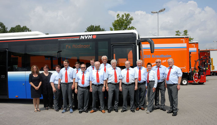 Vor verschiedenen LKW für Maschinentransporte ist ein blauer Bus für ein Gruppenfoto bereit gestellt. Davor sind 13 Herren und 2 Damen alle uniform in grau/blau/rot und die Damen in schwarz.