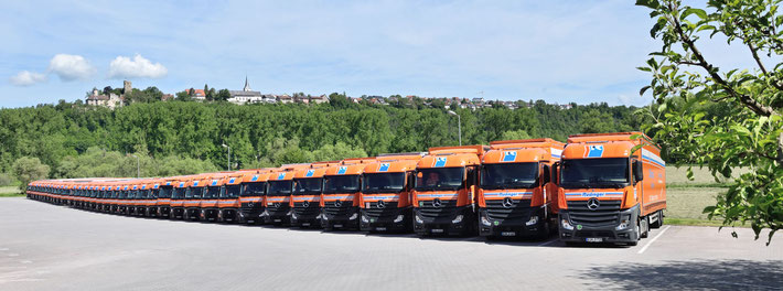 Zum Thema Maschinentrnasport sieht man auf eine Reihe von LKW einer Spedition. Es sind etwa 20 parallele vor blauem Himmel. Rechts hinten sind grüne Bäume