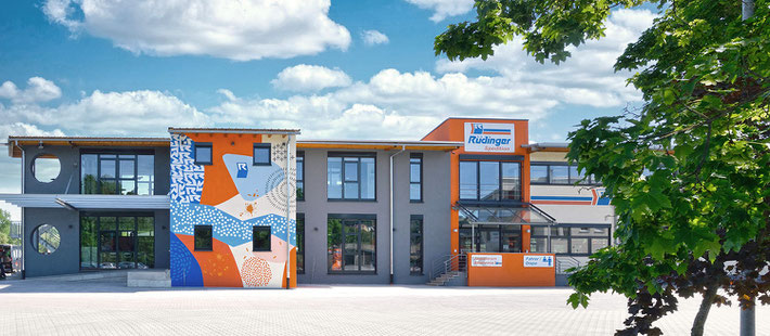 Man sieht links einen blauen Gelenkbus mit offenen Türen. Rechts daneben ist das neue Verwaltungsgebäude in orange/blau/weiß und rechts sieht man die Langguthalle mit vielen Menschen, die dort sitzen.