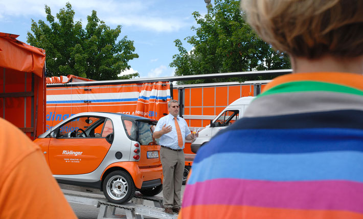 Der Inhaber der Spedition Rüdinger steht hinter dem Smart der Firma Rüdinger in orange, der gerade auf einen Maschinentransporter aufgeladen wird. Auffällig vorne rechts: ein tolles farbiges T-Shirt.