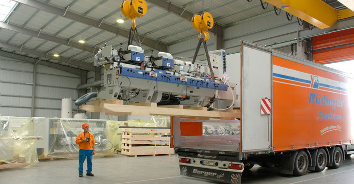 In großen Lettern steht Maschinentransport aus Leidenschaft. man sieht im Bild einen Mann mit Helm, wie er eine Maschine am Kran in ein LKW der Spedition Rüdinger navigiert.