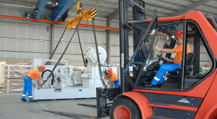 Oben steht Maschinentransport aus Leidenschaft in weißen Lettern. Man sieht die Verladung einer Maschine mit einem 12 Kran, links arbeiten 2 Mann an einer Maschine, rechts sitzt 1 Mann im Stapler.