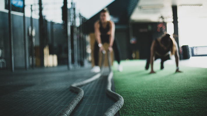 Auf dem Bild sind zwei Frauen im Fitnessstudio zu sehen. Das Motiv ist unscharf. Die linke Frau trainiert mit den Battleropes und die rechte Frau macht Liegestütze.