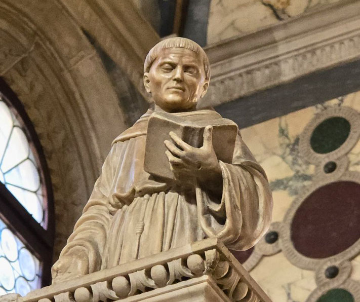 A detailed stone statue of a monk holding a book, set in an ornate interior with stained glass and marble decorations.