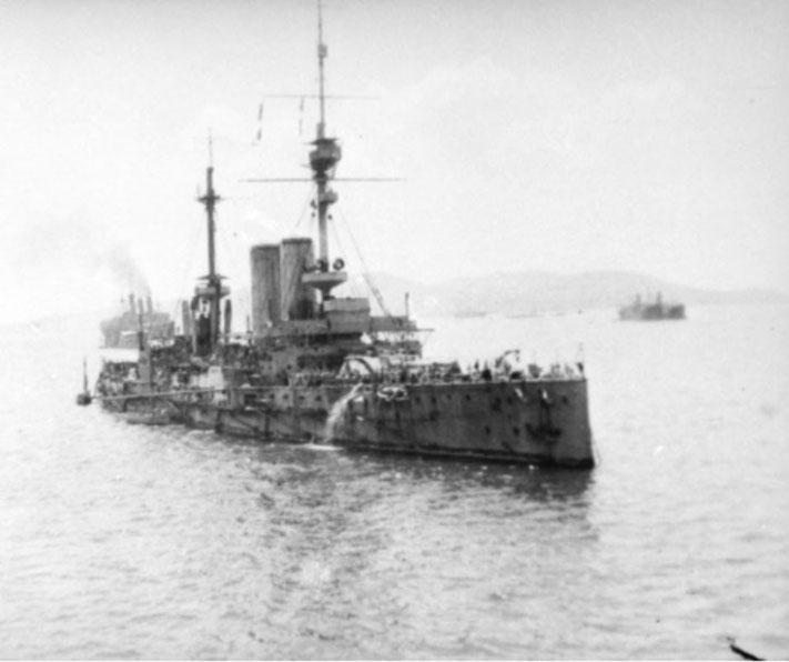 A battleship with twin smokestacks and tall masts sails in calm waters, crowded with crew on deck. Other ships are visible in the distance.
