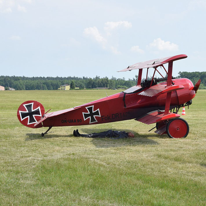 A red Fokker Dr.I triplane is parked on grass with a person lying underneath it, possibly for maintenance or shade.