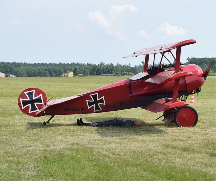 A red Fokker Dr.I triplane is parked on grass with a person lying underneath it, possibly for maintenance or shade.