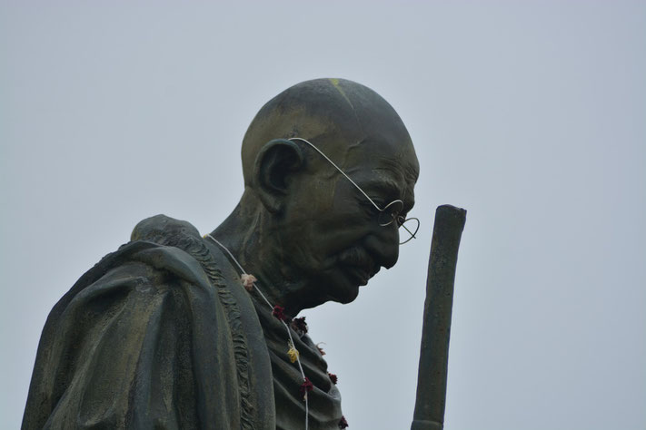 A bronze statue of Mahatma Gandhi shows him with glasses, staff, and a garland, gazing downward in contemplation.