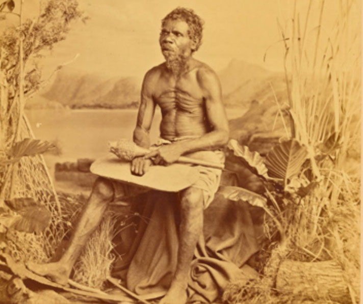 A historical sepia-toned photograph of an Indigenous Australian man sitting with a paddle, surrounded by vegetation.