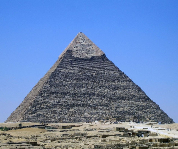 The Great Pyramid of Khufu at Giza, standing against a clear blue sky. Its weathered limestone blocks and partially intact casing stones at the top are visible, with visitors below.
