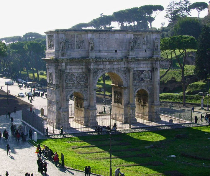 Triumphal Roman arch surrounded by visitors and greenery, with sunlit trees and ancient pathways in the background.