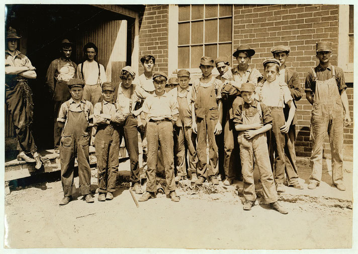A group of young boys and men in worn overalls and caps stand outside a brick factory.