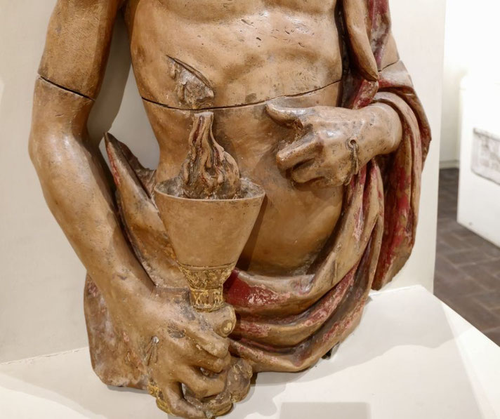 Wooden torso sculpture of a robed figure holding a flaming chalice, with a visible wound on the chest and detailed hand positioning.