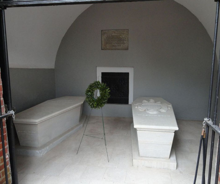 Two white marble tombs inside a gated crypt with a wreath placed between them, set beneath a vaulted ceiling and commemorative plaque.