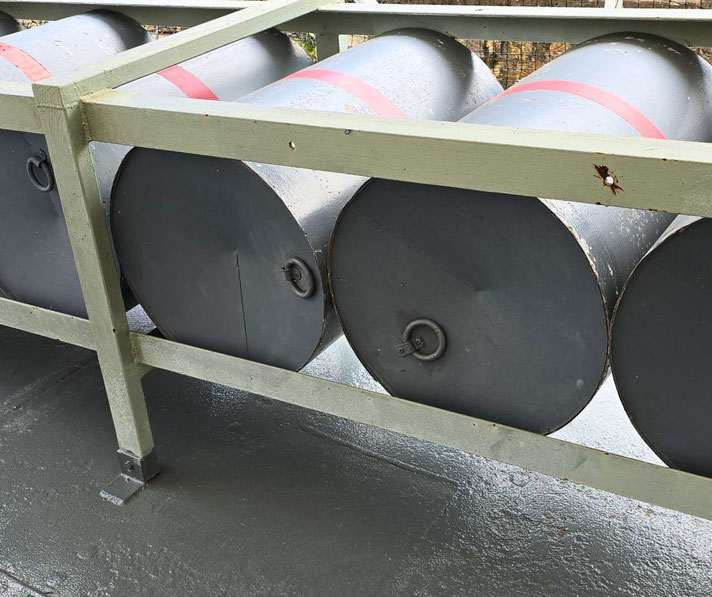 Four dark cylindrical depth charges with red bands are secured in a green metal rack on a ship’s deck.