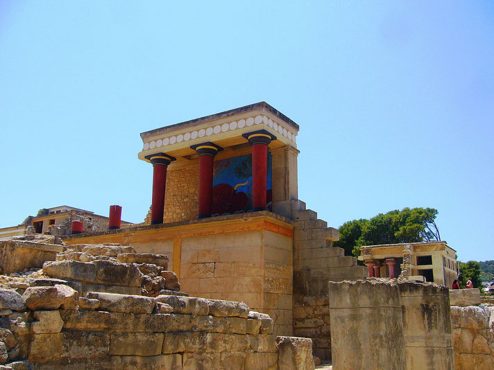 Colorful ruins of the Palace of Knossos featuring red columns, stone walls, and a fresco under a clear blue sky.