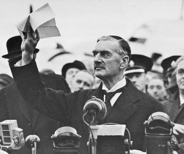 A man in a suit raises a document while speaking into multiple microphones, surrounded by a crowd.