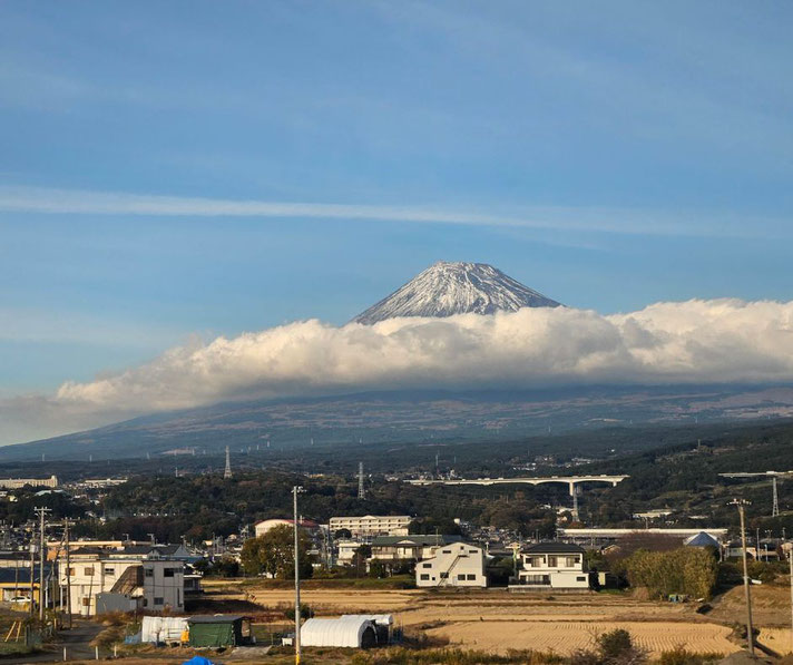 Mount Fuji with its snow-capped peak rising above a layer of clouds. Below, a rural landscape with houses, fields, and infrastructure stretches under a clear blue sky.