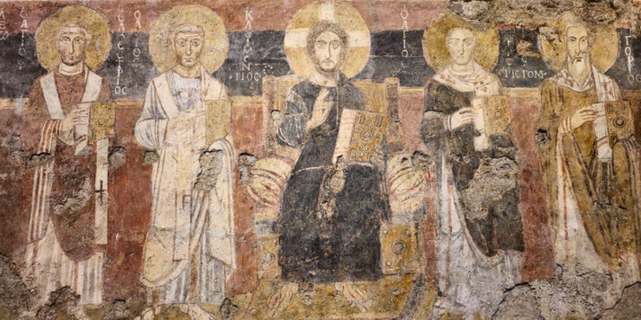 Faded fresco of five haloed religious figures holding books, with central figure likely Christ, seated and flanked by saints, on a worn and partially damaged wall.