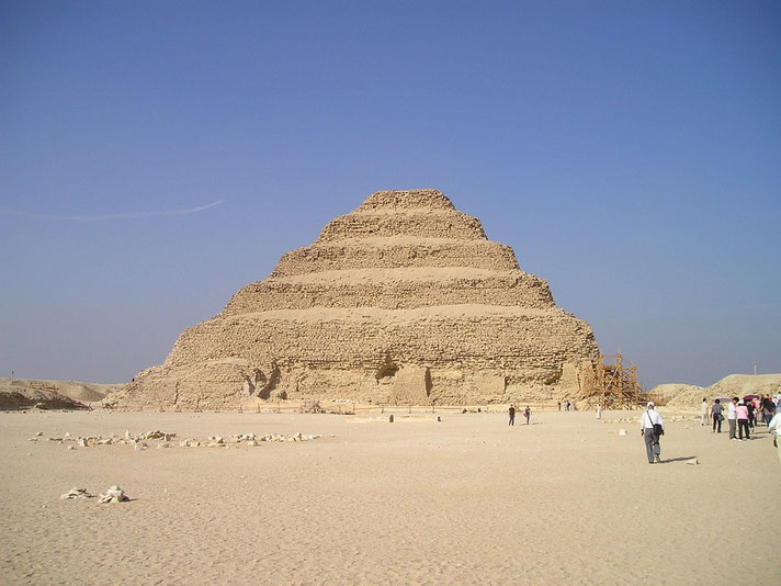Step Pyramid of Djoser at Saqqara, Egypt, an early example of monumental stone architecture, standing in a vast desert with visitors walking nearby under a clear blue sky.