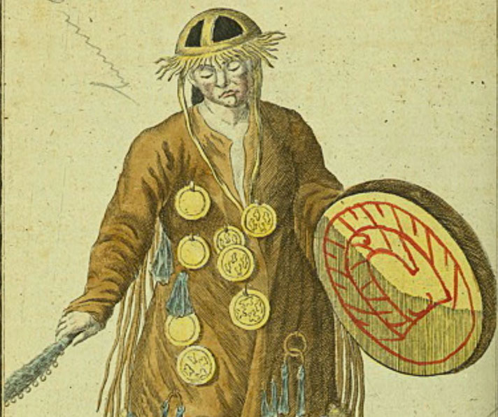 An old illustration of a shaman wearing a brown robe adorned with golden medallions, a fringed hat, and holding a decorated drum with red symbols. He carries a ritual object in his other hand.