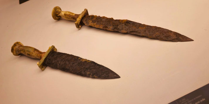 Two ancient double-edged daggers with corroded iron blades and short, rounded hilts, likely bronze or wood, showing heavy rust and age.