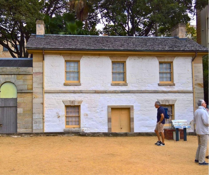A historic white stone cottage with a shingled roof, wooden window frames, and a small door stands among trees. 