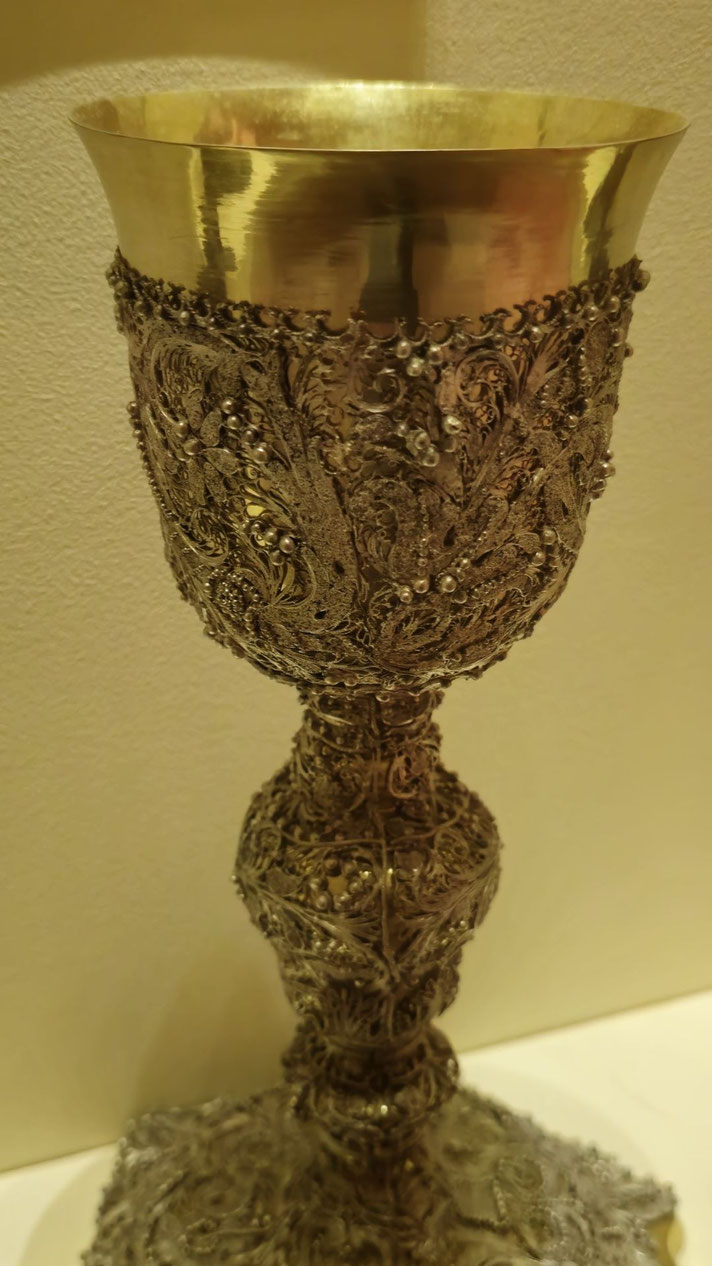 Gold and silver chalice with a smooth rim and heavily detailed body covered in floral patterns, filigree, and small pearl-like accents.